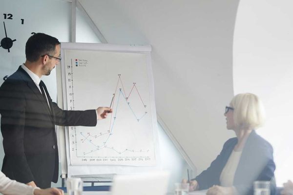 A man gives a presentation to a group of people. His chart shows a graph trending upwards.